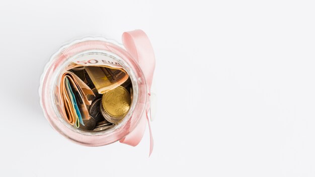 Euro notes and coins in glass jar tied with pink ribbon on white background