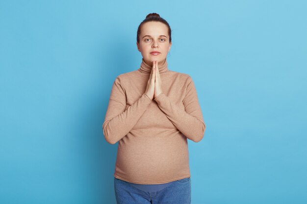 European beautiful pregnant woman with palms together praying for her future baby wearing casual attire, stands isolated on blue wall, tries to relax before going to maternity house.