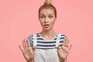 Free photo european blonde female poses with outstretched hands, demonstrates stop gesture, asks to halt, has surprised scared expression, demands not bother her, shows prohibition, wears fashionable overalls