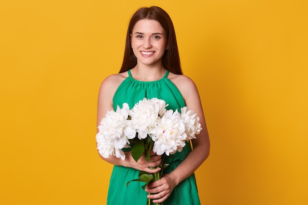 Free photo european brunette woman wearing green sundress holding bouquet of white peonies flowers in both hands, posing isolated on yellow, being in high spirits. sping concept.