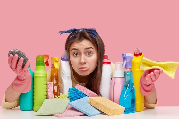 European clueless young housekeeper looks with apathy at camera, spreads palms, wears protective gloves, carries rag