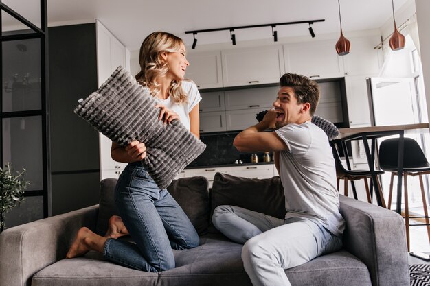 European couple posing during pillow fight. Indoor portrait of laughing young people chilling in living room.