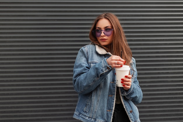 Европейская милая молодая хипстерская женщина в модной джинсовой синей куртке в модных фиолетовых очках с чашкой горячего кофе стоит возле серой стены в городе. довольно красивая девушка позирует на улице.