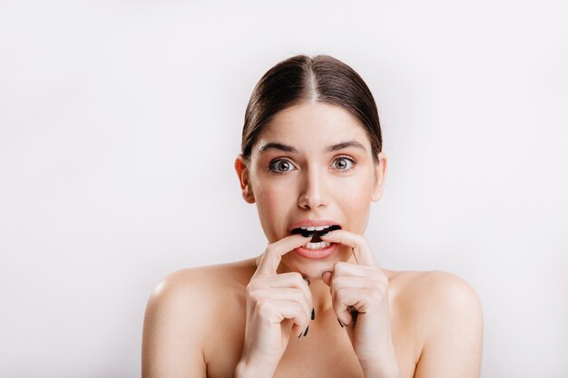 European female model with healthy skin bites her fingers for fear, posing on isolated wall.