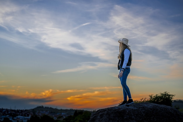 岩の上に立って夕日を見ているカウボーイハットを持つヨーロッパの女性