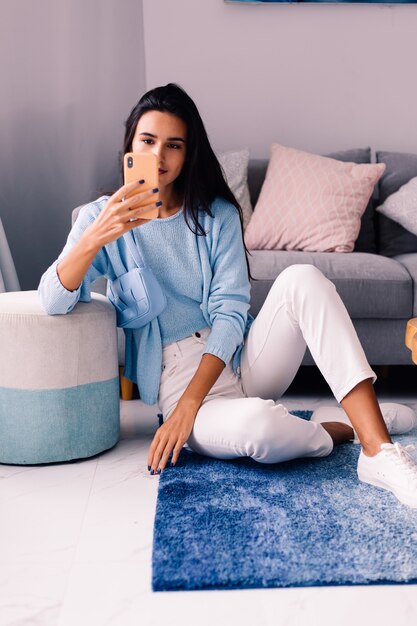 European fit brunette fashion blogger woman sits on floor in living room near sofa with phone