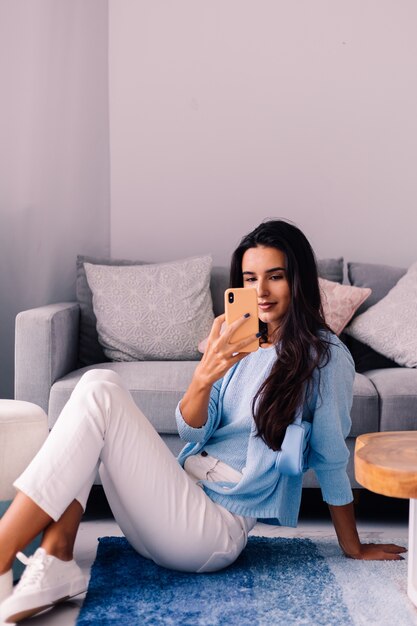 European fit brunette fashion blogger woman sits on floor in living room near sofa with phone