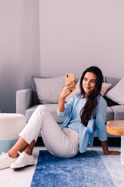 European fit brunette fashion blogger woman sits on floor in living room near sofa with phone