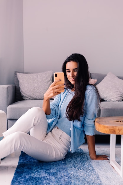 European fit brunette fashion blogger woman sits on floor in living room near sofa with phone