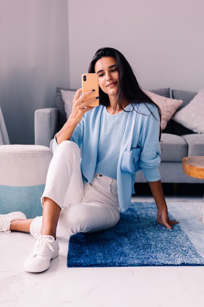 European fit brunette fashion blogger woman sits on floor in living room near sofa with phone