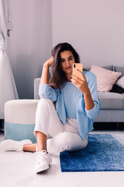 Европейская подтянутая брюнетка модный блоггер сидит на полу в гостиной возле дивана с телефоном