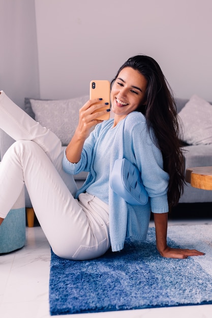 European fit brunette fashion blogger woman sits on floor in living room near sofa with phone