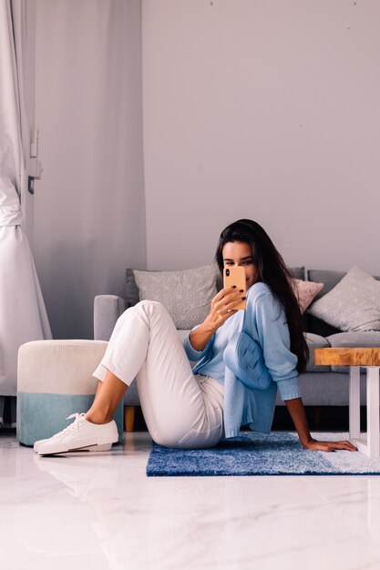 European fit brunette fashion blogger woman sits on floor in living room near sofa with phone