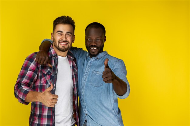 European guy and afroamerican guy are laughing and looking in front of them with thumbs up in informal clothes 