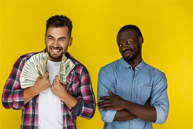 European guy with big amount of money in both hands is hapily smiling and afroamerican guy is having nothing is looking on him in informal clothes 