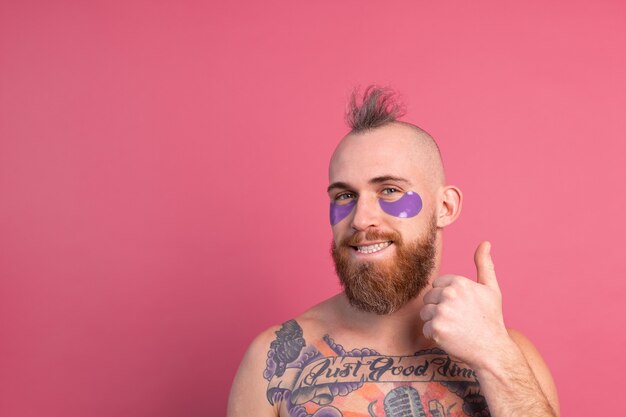 ピンクのカメラにポーズをとる紫色の眼帯マスクを持つヨーロッパのハンサムなひげを生やした入れ墨トップレスの男