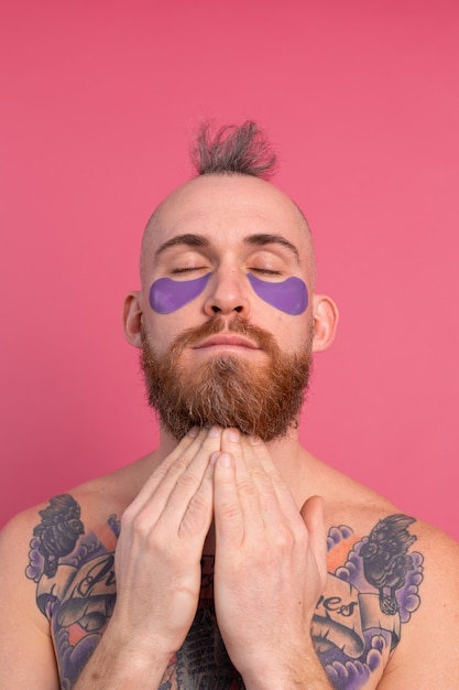 European handsome bearded tattooed topless man with purple eye patches mask posing to camera on pink