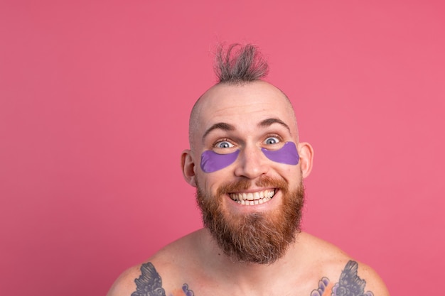 European handsome bearded tattooed topless man with purple eye patches mask posing to camera on pink