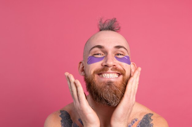 European handsome bearded tattooed topless man with purple eye patches mask posing to camera on pink