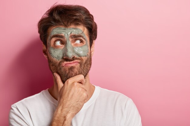 ヨーロッパ人の男性は、あごを持ち、唇をすぼめ、思いやりのある表情、太い毛、脇を向いて、顔に粘土のマスクを適用し、白いTシャツを着て肌をリフレッシュし、ピンクの壁にモデルを置きます。男性的な美容
