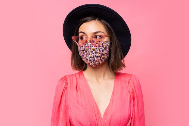 Maschera facciale elegante protettiva vestita modello europeo. indossare cappello nero e occhiali da sole. in posa sul muro rosa