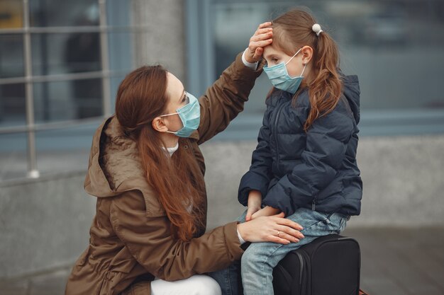 A European mother in a respirator with her daughter are standing near a building.The parent is teaching her child how to wear protective mask to save herself from virus