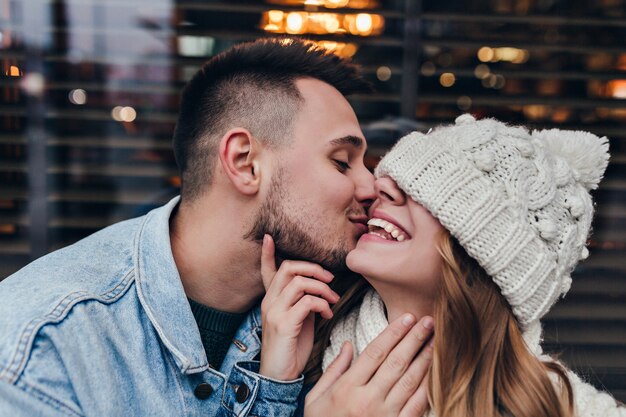 european pretty girl in hat laughing while boyfriend kissing her. outdoor photo of blissful brunette guy having fun with girlfriend on urban street.