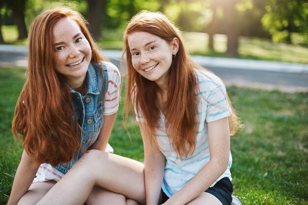 european sisters with red hair and freckles sitting on green grass and smiling broadly, hanging out with friends on picnic, expressing joy and amusement. Emotions and family concept