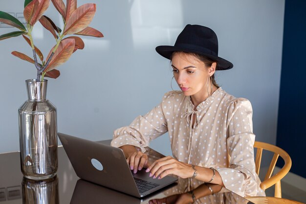 European woman in dress and classic hat works at home in kitchen