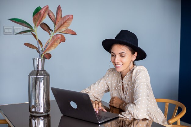 European woman in dress and classic hat works at home in kitchen