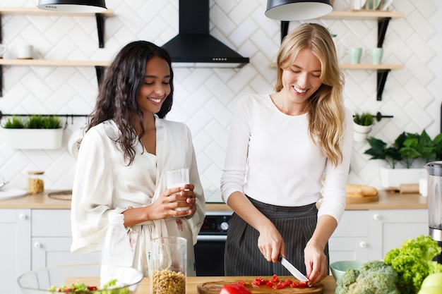 European woman is cutting a tomato and african woman is drinking milk, they smile