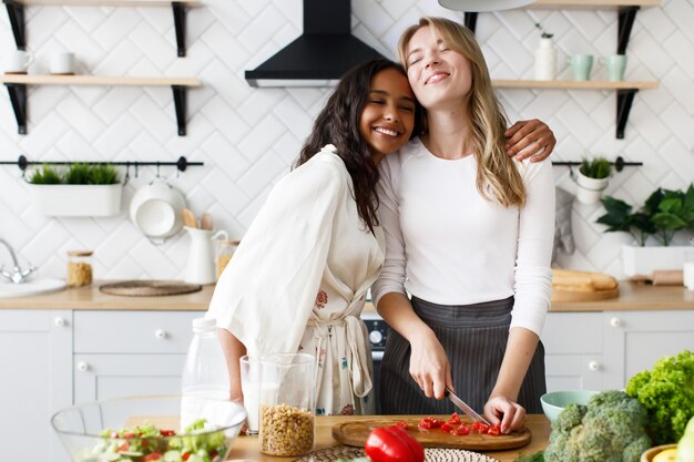 European woman is cutting a tomato and african woman is hugging her, they smile