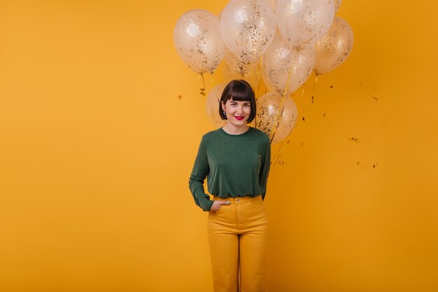 European woman posing with shy smile. Indoor portrait of cheerful lady standing with hand in pocket.