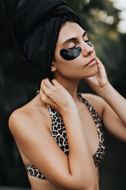 European woman in turban posing with closed eyes on nature background. Outdoor shot of relaxed lady in swimsuit.