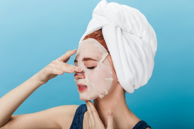 European woman with towel on head applying face mask. Studio shot of amazing girl doing spa treatment on blue background.