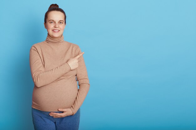 European young pretty pregnant woman touching belly with one hand and pointing aside with index finger, standing isolated on blue wall,  expectant mother with hair bun.