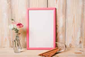 Free photo eustoma in glass vase; colored pencils and white picture frame with pink border on wooden table