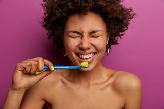 日常生活、朝のルーチン、歯のクリーニングのコンセプト。上半身裸の暗い肌の女性の水平方向のショットは、歯ブラシで歯を磨き、紫色の壁に裸で立って、機嫌が良い