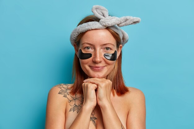 Everyday routine. Beautiful redhead woman keeps hands under chin, wears collagen patches under eyes, has regular rejuvenation treatment for healthy skin, wears bow hairband, stands bare shoulders