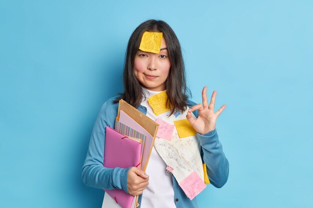 Everything is under control. Serious self confident young Asian woman makes okay gesture agrees to collaborate with colleague for preparing research work surrounded with papers and stickers.