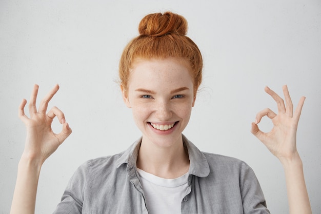 Everything is just fine! Cheerful excited young Caucasian female with ginger hair knot and freckled skin showing Ok gesture with both hands and smiling broadly, enjoying her carefree happy life