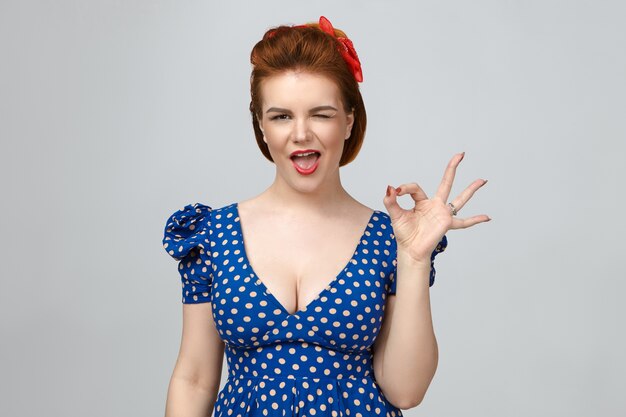 Everything is perfect. Isolated picture of elegant gorgeous young ginger female wearing red lipstick and vintage blue dress blinking at camera, keeping mouth wide opened, showing ok gesture