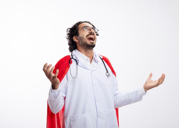 Free photo evil joyful young caucasian superhero man in optical glasses wearing doctor uniform with red cloak and with stethoscope around neck holds hands open and looks up isolated on white wall