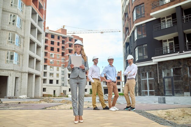 examination. serious young adult woman in business gray suit in heels wearing safety helmet looking at document and smiling three men waiting behind at construction site