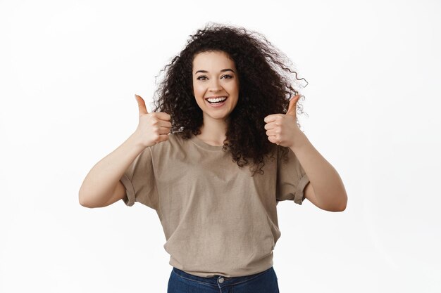 Excellent choice. Brunette girl shows thumbs up, smiles and looks satisfied, pleased by good result, praise great work, excellent job, standing over white background.