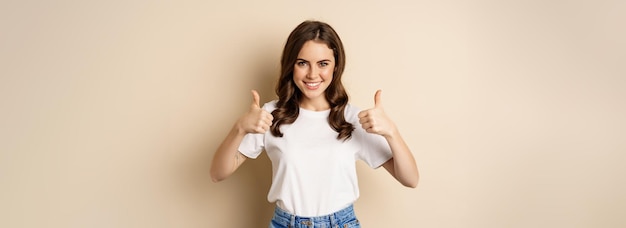 Excellent work happy young woman showing thumbs up in approval and smiling standing in tshrit over b