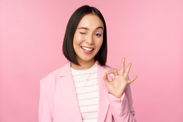 훌륭한 일을 잘한 제스처 웃는 아시아 여성 사업가가 OK 표시를 하고 자신감 있는 전문 분홍색 배경으로 보이는 회사를 추천하는 좋은 일을 칭찬합니다.