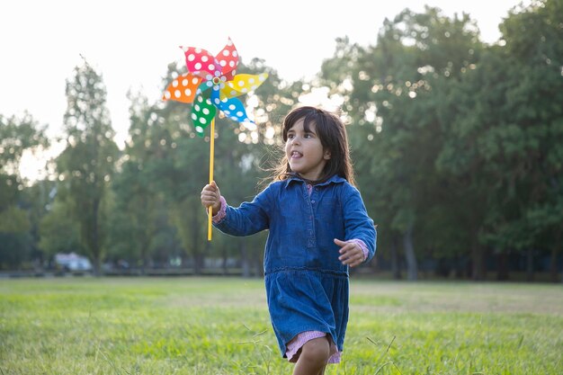 Excited adorable black haired girl holding pinwheel and running on grass in park. Children outdoor activity concept