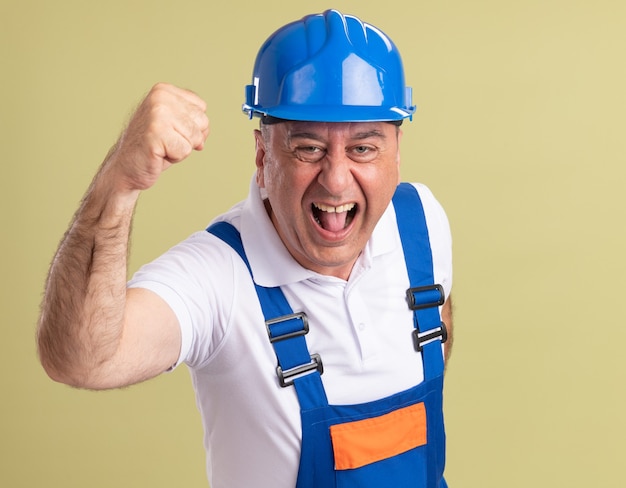 Uomo adulto emozionante del costruttore in uniforme che alza il pugno su verde oliva