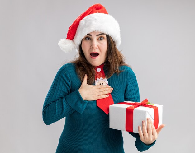 excited adult caucasian woman with santa hat and santa tie puts hand on chest and holds christmas gift box isolated on white background with copy space